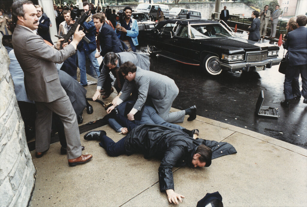 Secret Service Agents cover Press Secretary James Brady moments after he is shot while a secret service agent holds an UZI submachine gun in case there are further attacks.