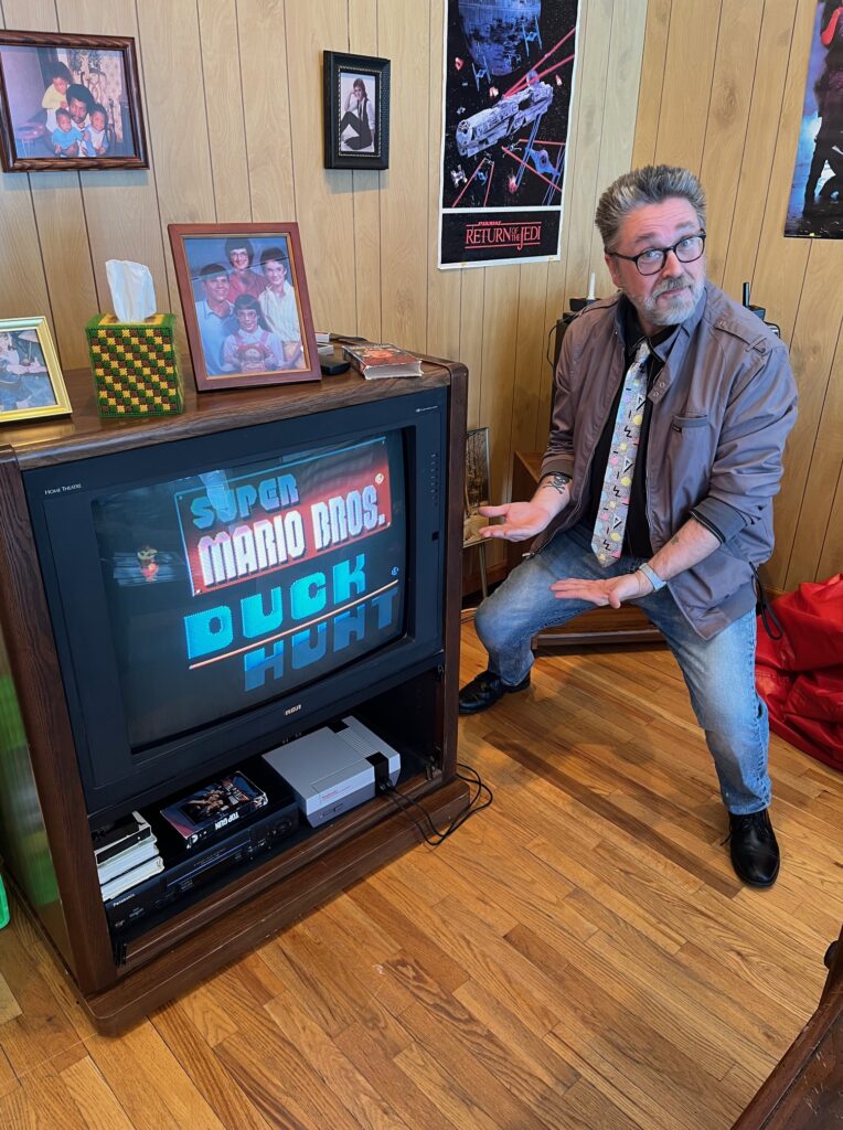 Me standing by a large TV with the NES system duck hunt game on the television.