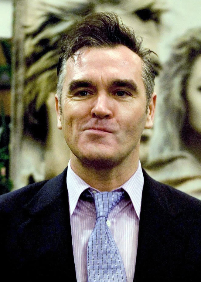 Morrisey sneering into the camera wearing a black suit, blue tie, and white dress shirt. 