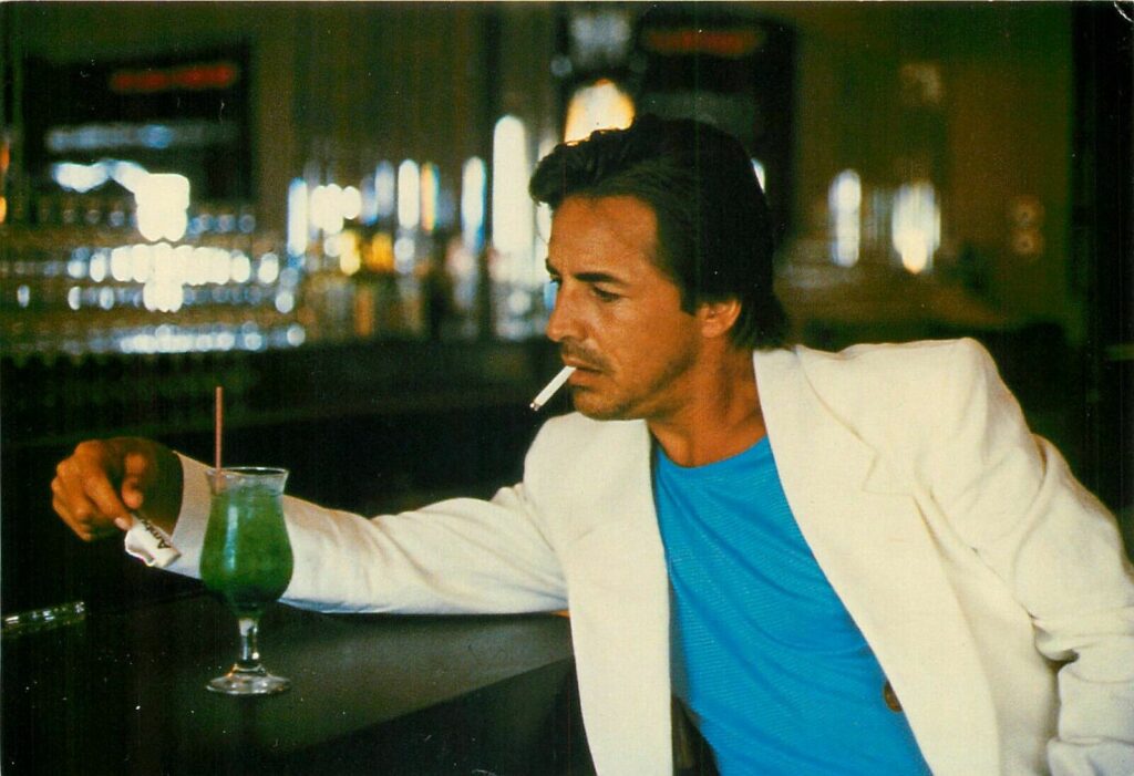Don Johnson in white suit with shoulderpads and blue t shirt smoking a cigarette leaning against a bar by a tropical drink.