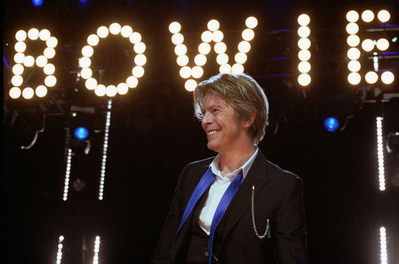David Bowie smiling wearing a black suit and a loosened blue tire below lights that read 'BOWIE'