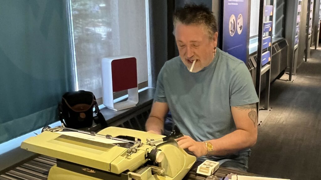 Man types on a yellow typewriter while smoking a cigarette. 