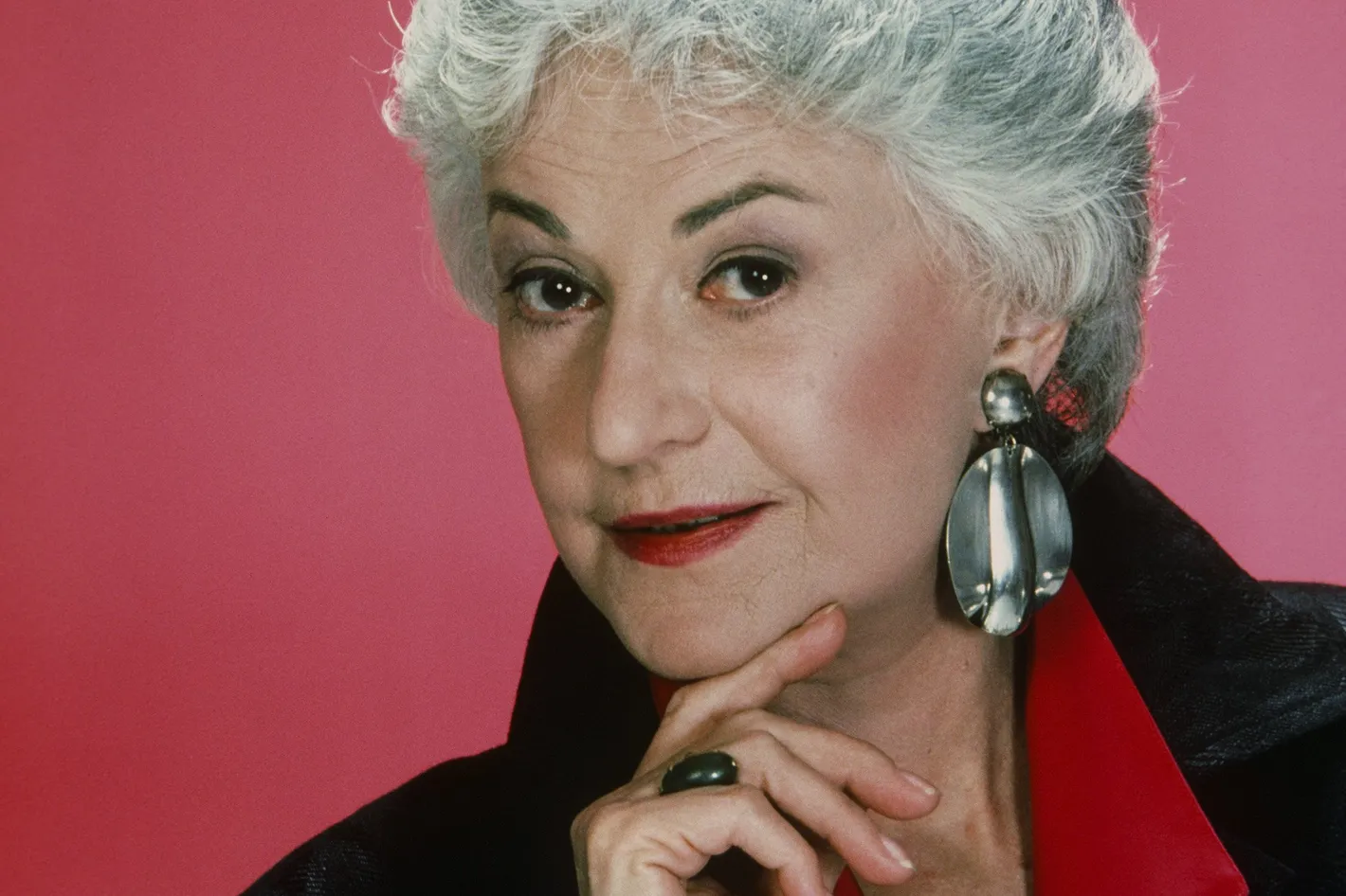 Bea Arthur with gray hair, fashionable black and red jacker and silver earrings.