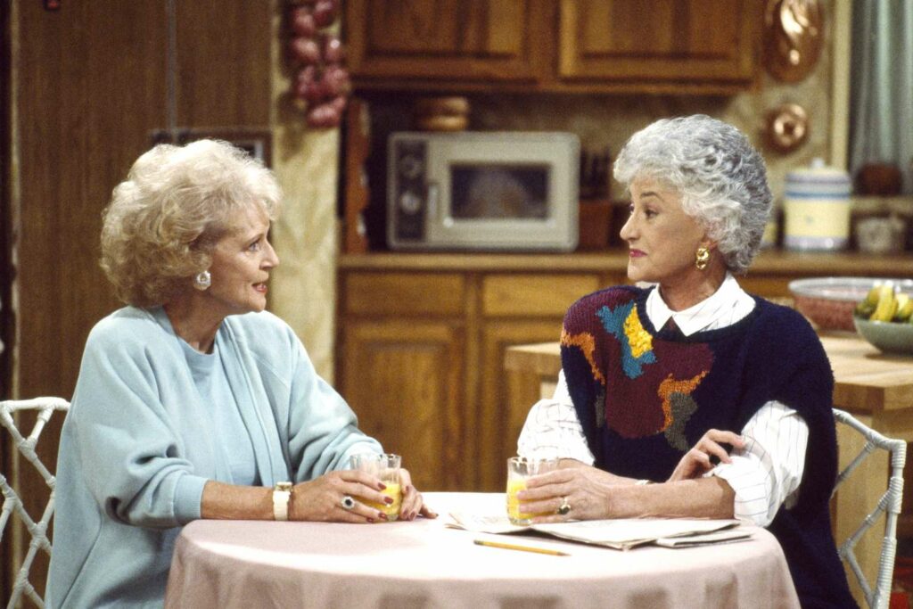 Still of Bea Arthur and Bette White from Golden Girls sitting at a table drinking juice and talking.