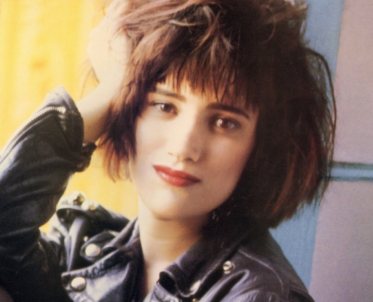 Martika with short black hair, leather jacket, red lipstick running a hand through her hair.