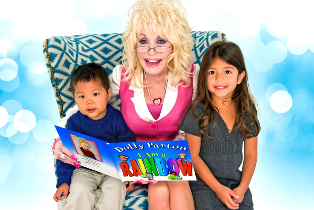 Dolly Parton in a pink dress wearing reading glasses while on a love seat in the clouds with a child sitting on either side to them as she holds a book. 