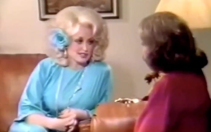 Dolly Parton in a blode wid, blur dress with a blur bow. The back of Barbara Walters is in the foreground. Dolly is leaning in confidently.