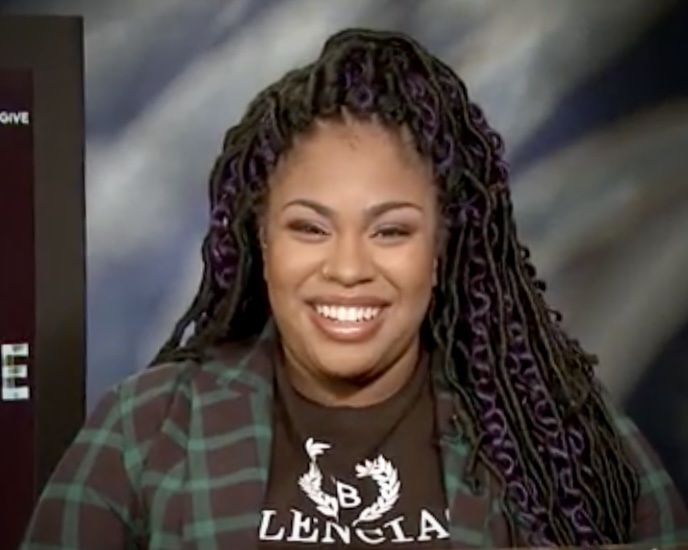 Angie Thomas. A black woman with long hair and a flannel shirt smiling.