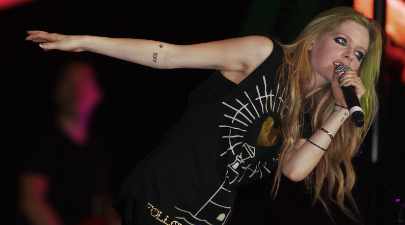 Avril Lavigne leaning right into a microphone with her arm outstretched left in a powerful stage pose.