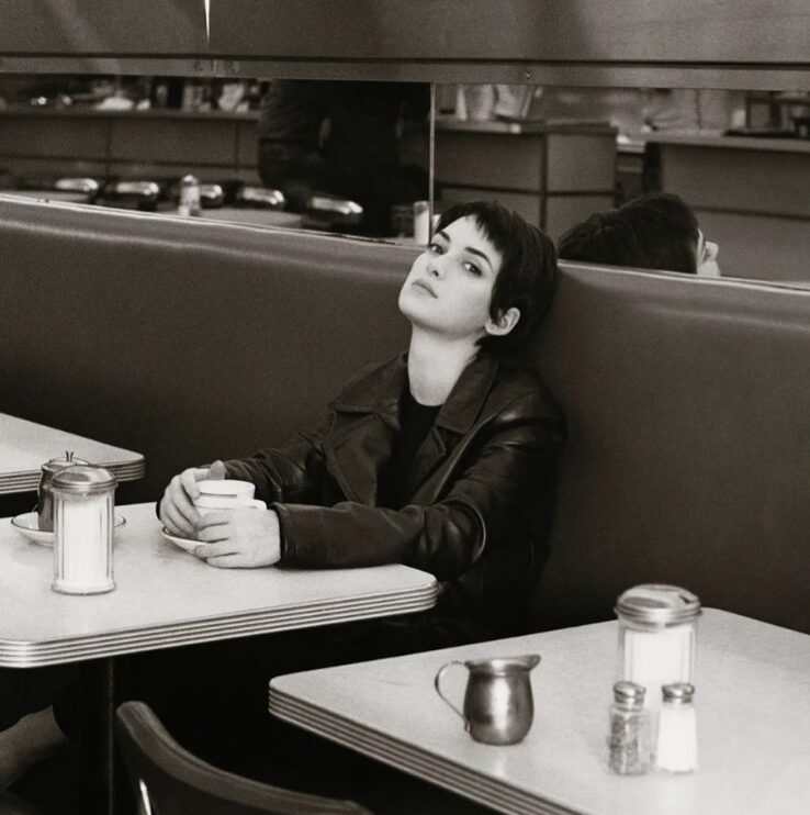 Winona Ryder with pixie short hair holding a cup of coffee in a late night diner