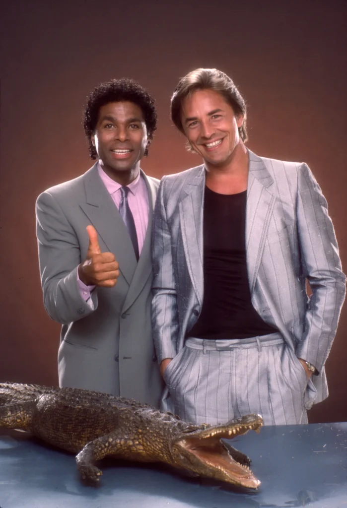 Black man in grey suit and white man in silver suit with black t shirt by a plastic alligator