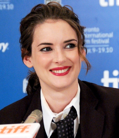 Winona Ryder smiling while wearing a black jacket and tie near a microphone.