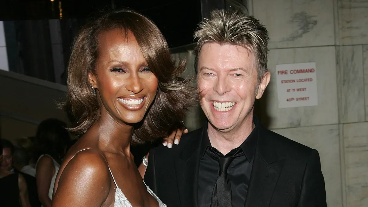 Iman in a white dress next to David Bowie in a black suit. Both are smiling.