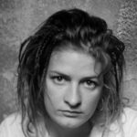 Black and white photo of young woman staring directly at camera with dark hair and a white shirt and serious face.