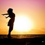 Silhouette of a woman at the beach with arms outstretched against a vibrant sunset backdrop.