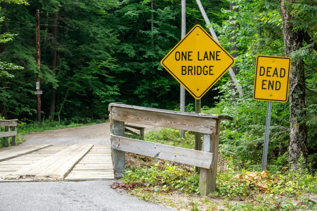 A rustic wooden one-lane bridge with 'Dead End' and 'One Lane Bridge' signs in a lush forest setting.